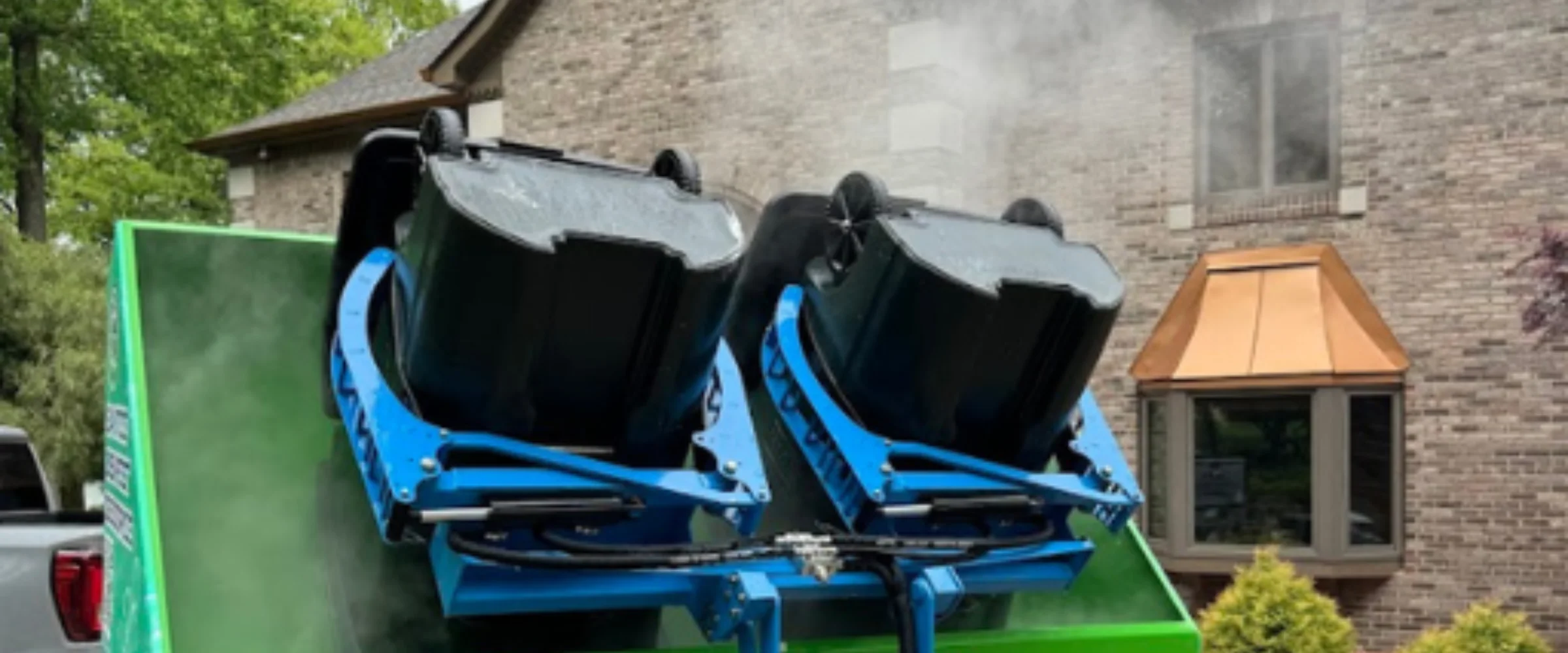 Two black trash bins are inverted in a blue and green cleaning system, emitting steam against a brick house background. Two black trash bins are inverted in a blue and green cleaning system, emitting steam against a brick house background.