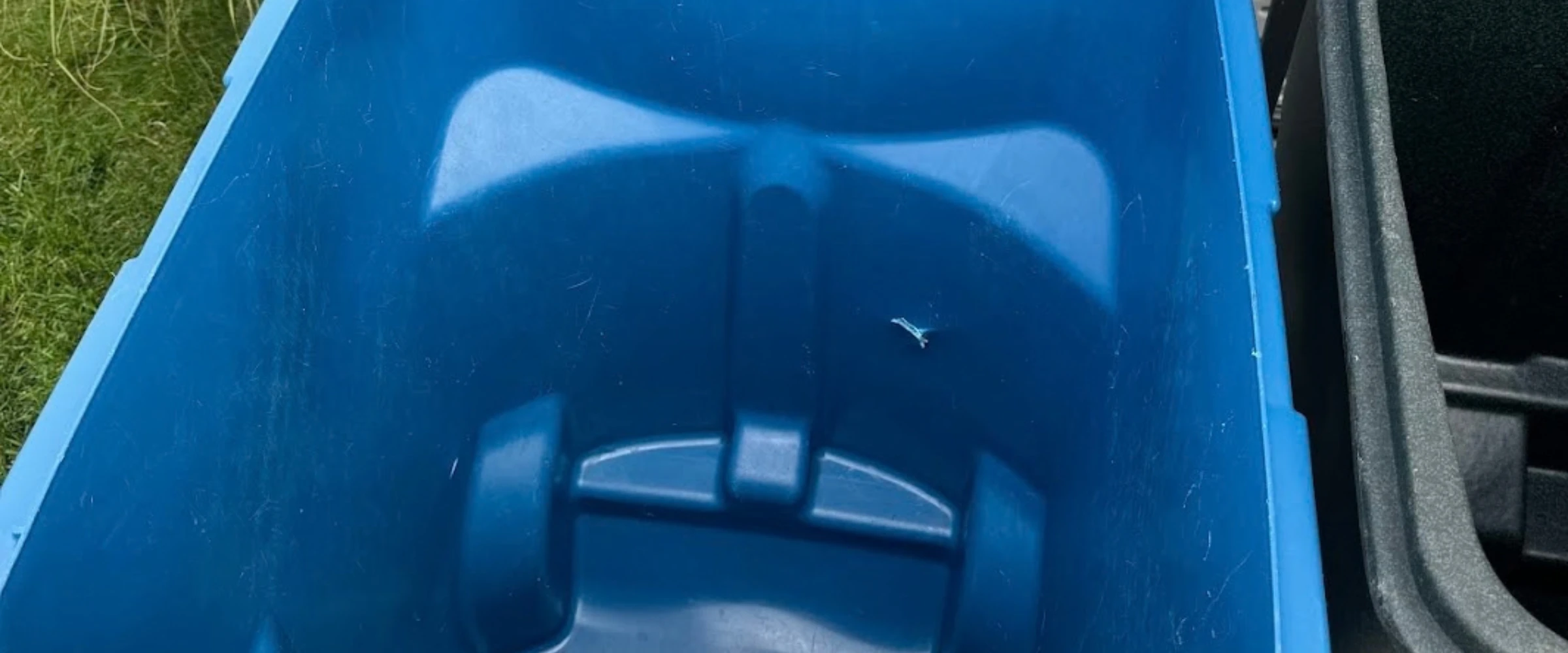Close-up view of the interior of an empty blue plastic recycling bin, with another darker bin and green grass visible. Close-up view of the interior of an empty blue plastic recycling bin, with another darker bin and green grass visible.