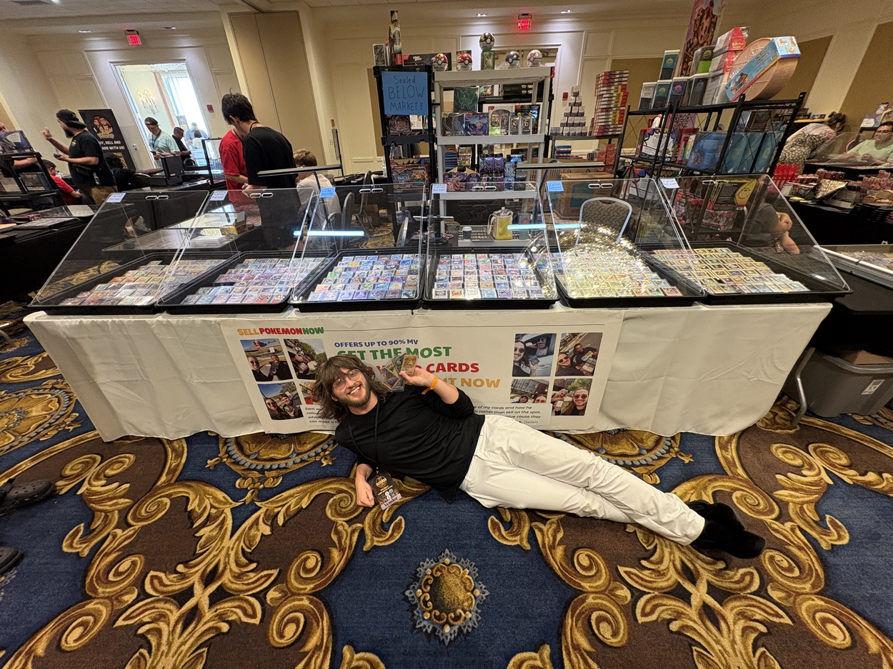 Sam in front of his table at a card show, with binders and display cases of Pokémon cards