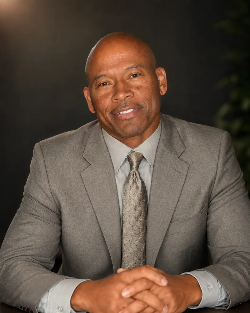 Bill Saulsberry, founder of Mustard Seed Concepts, seated at a table in a grey suit, smiling, hands clasped