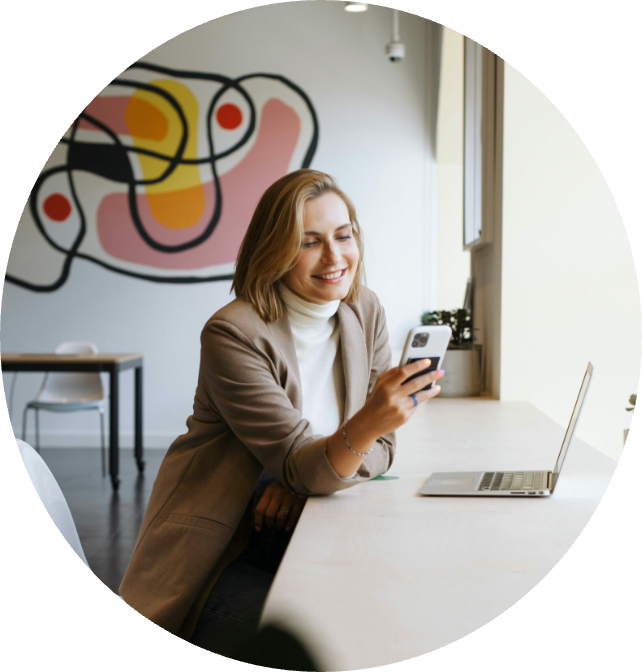 Smiling woman in a modern workspace using smartphone, with a laptop on the table and abstract wall art in the background, representing virtual assistant services and productivity.