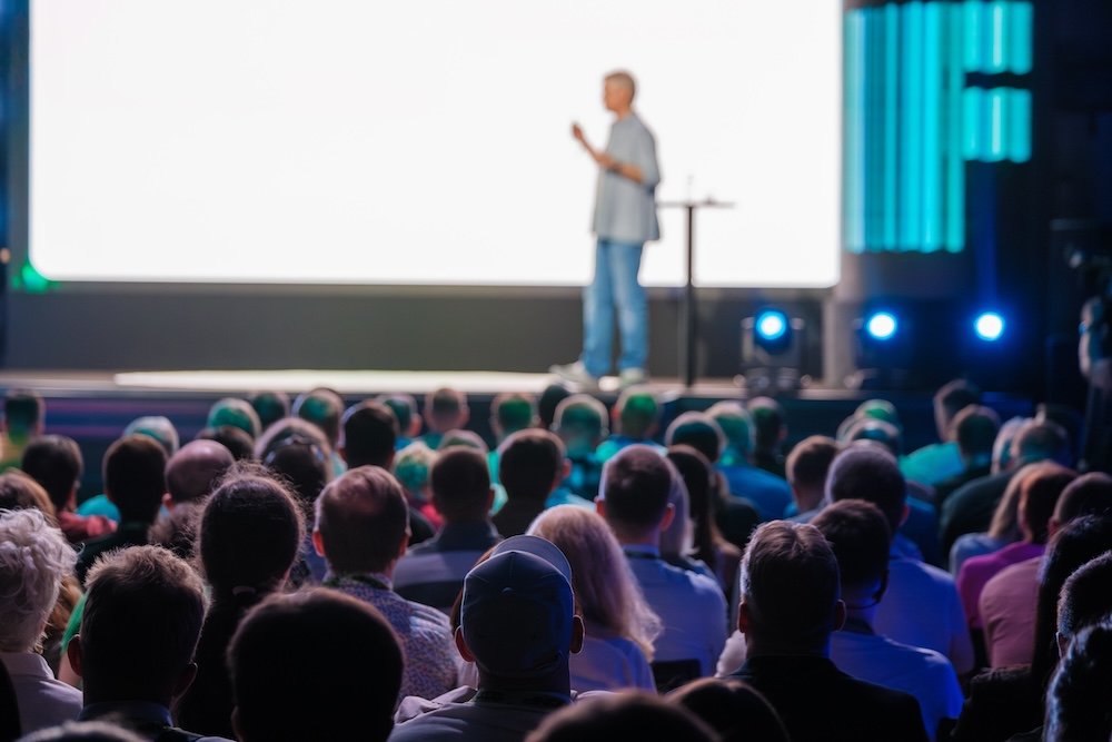 Audience watching a speaker on stage, with a blank presentation screen behind, highlighting the Speaker Support Services offered by Every Task for effective engagement management.