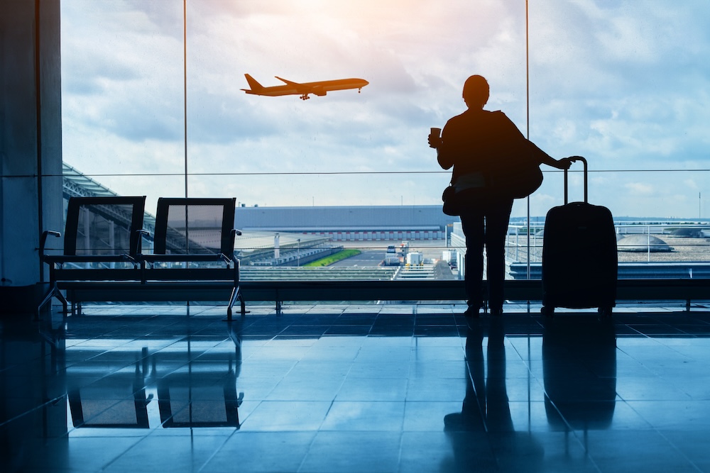 Silhouette of a traveler at an airport window, holding a coffee cup and a suitcase, with a plane taking off in the background, symbolizing seamless travel arrangements.