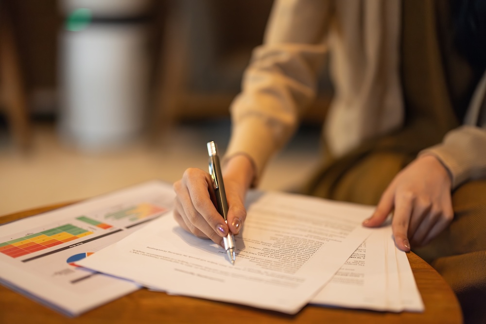Person writing on grant proposals with charts and documents on a table, illustrating the grant writing process for funding opportunities.