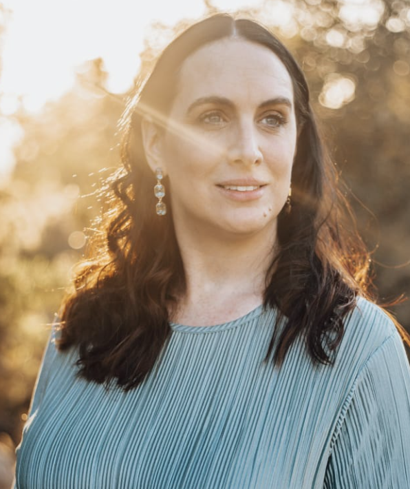 Woman with long dark hair wearing a pleated light blue top and earrings, standing outdoors with sunlight shining through trees, conveying a sense of confidence and professionalism related to customer service support.