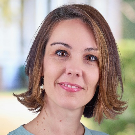 Professional woman with shoulder-length hair, smiling, in a soft-colored top, set against a blurred outdoor background, representing a virtual assistant instructor for online courses.