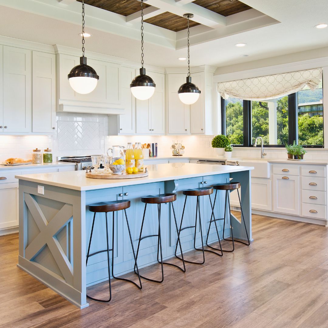 Bright farmhouse kitchen with a blue island, white shaker cabinets, pendant lighting, and large windows by Elci Cabinets and Floors.