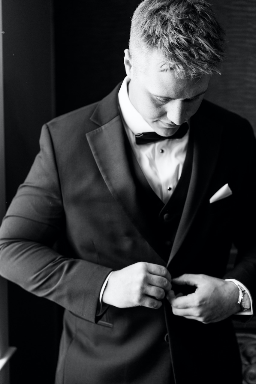Black-and-white portrait of a groom in a tuxedo and bow tie buttoning his jacket as he gets ready