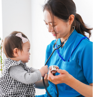 A happy nurse in blue scrubs examining a cute baby girl dressed in a checkered dress with a light pink bow in her hair.