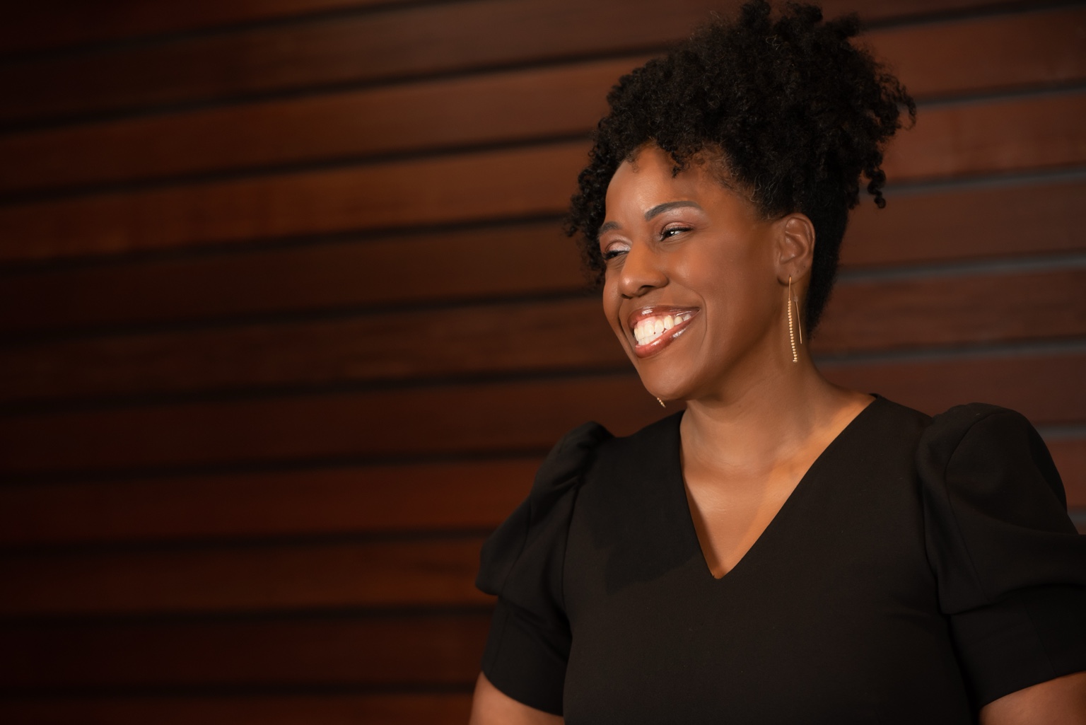 Aesha Ray smiling in a black dress against a warm wood backdrop