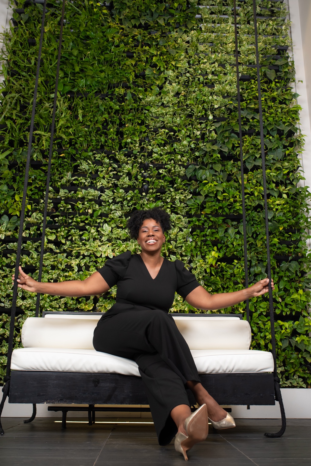 Aesha Ray smiling on a swing in front of a green living wall