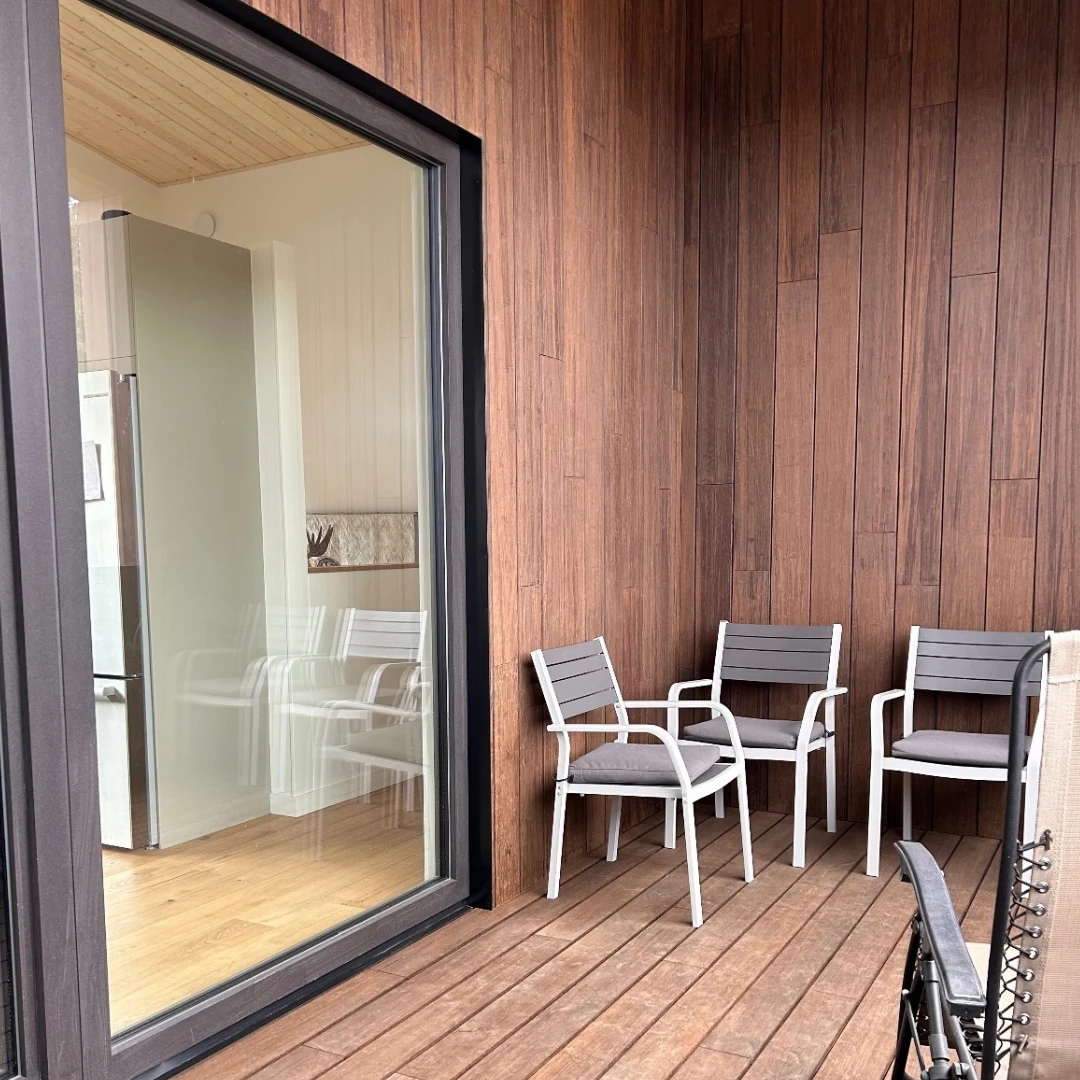 Outdoor patio area featuring vertical wood siding, wooden deck floor, and white patio chairs near a sliding glass door.