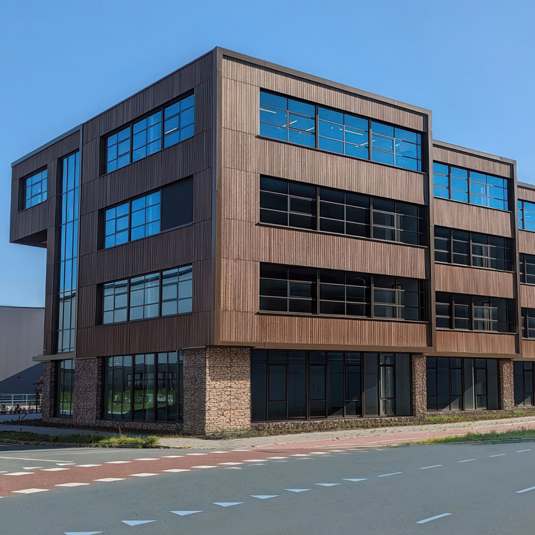Wide shot of a modern, multi-story commercial building with brown, wood-like paneled facade and large glass windows. Ground floor features stone cladding. A paved road is in the foreground. Clear blue sky.