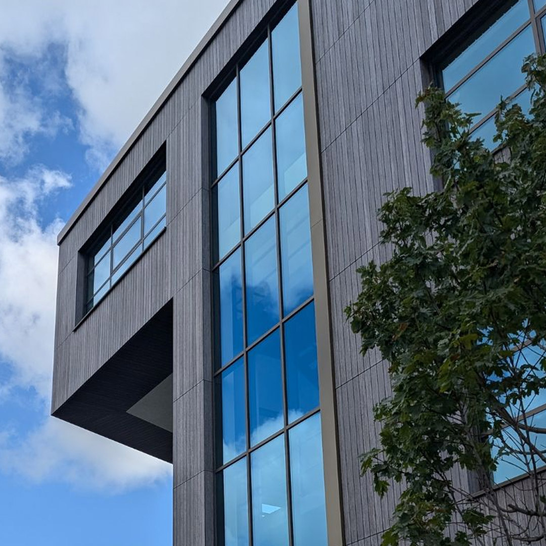 Close-up angled shot looking up at the facade of a modern commercial building with gray textured panels and large blue-tinted windows reflecting the sky and clouds. A tree branch is visible on the right. Blue sky with white clouds in the background.