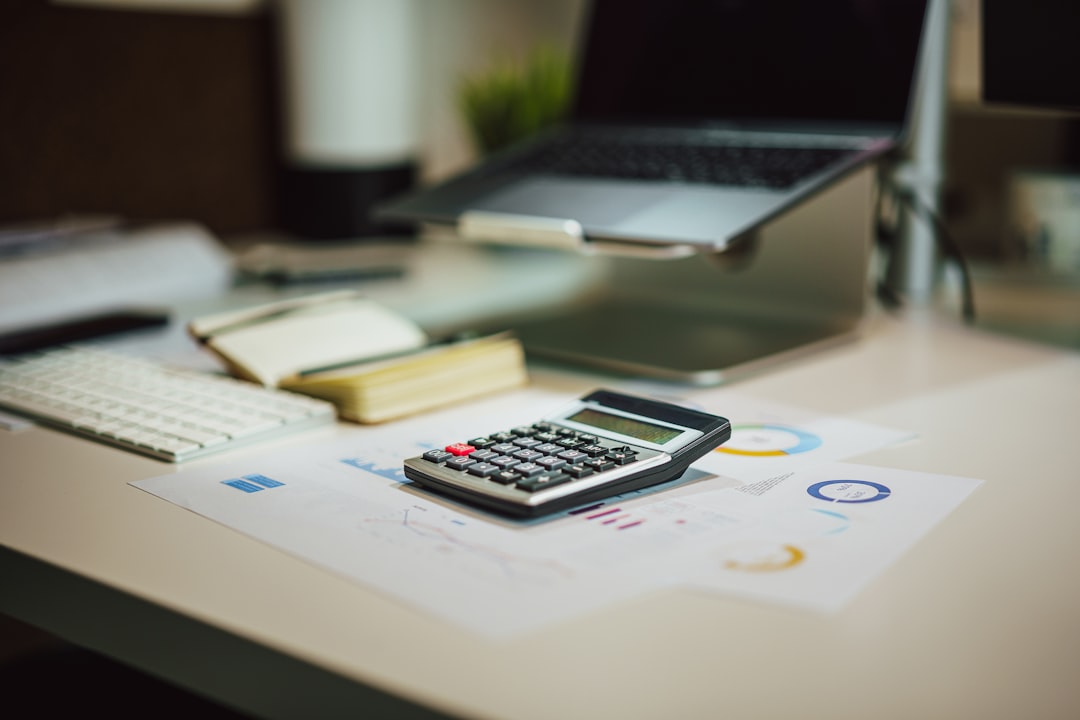 Calculator resting on printed charts on a white desk beside a laptop on a stand, keyboard and notepad nearby.