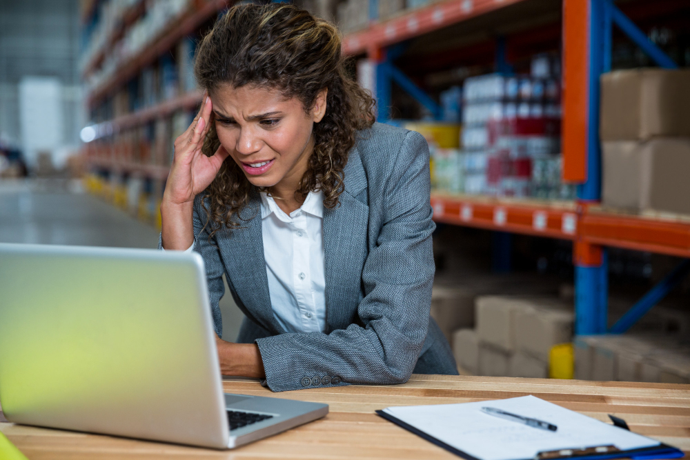 Woman in a warehouse office looks stressed while working on a laptop, holding her temple with one hand; shelves of boxed inventory are visible in the background. Woman in a warehouse office looks stressed while working on a laptop, holding her temple with one hand; shelves of boxed inventory are visible in the background.
