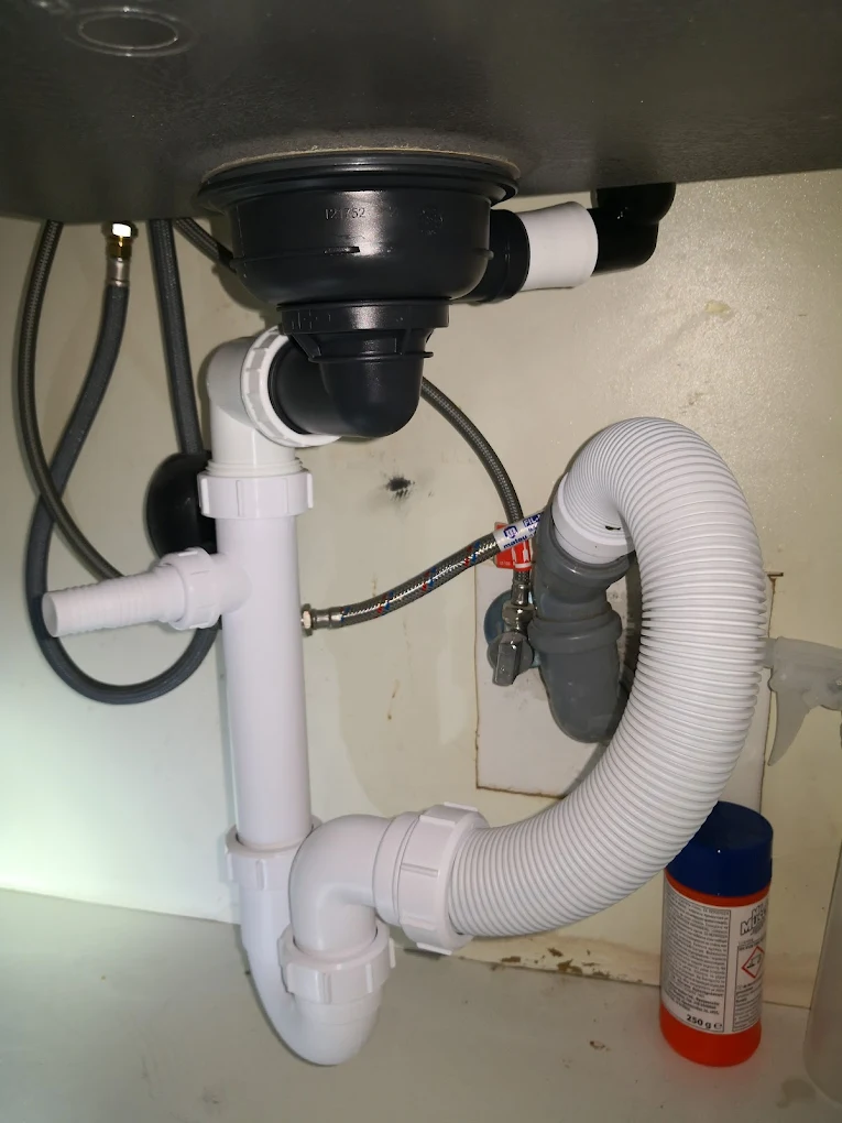 Mosogatótálca alatti szerelvények sűrűje egy szekrény mélyén. A fekete lefolyóhoz egy összetett, fehér műanyagból épített szifonrendszer csatlakozik, amelyen egy oldalra kinyúló, bordázott végű bekötőcsonk is megfigyelhető. A szennyvíz egy vastag, harmonikaszerűen hajlítható, U-alakban felkanyarodó gégecsövön keresztül távozik a fali szürke csatornakiállásba. A csőrengeteg mögött vékony, fémhálós vízvezetékek tekeregnek, a jobb alsó sarokban pedig egy élénk narancs-kék flakonos tisztítószer bújik meg.