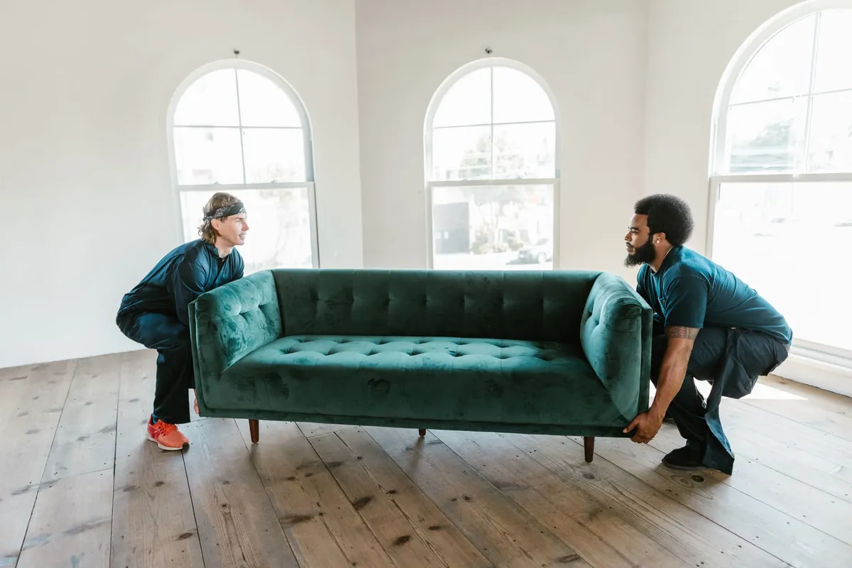 Two movers carrying a green sofa through a bright room with arched windows