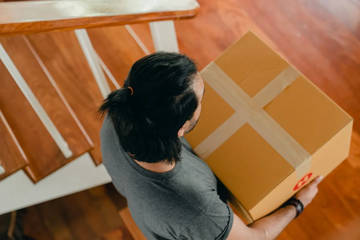 Person carrying a sealed cardboard box down a wooden staircase