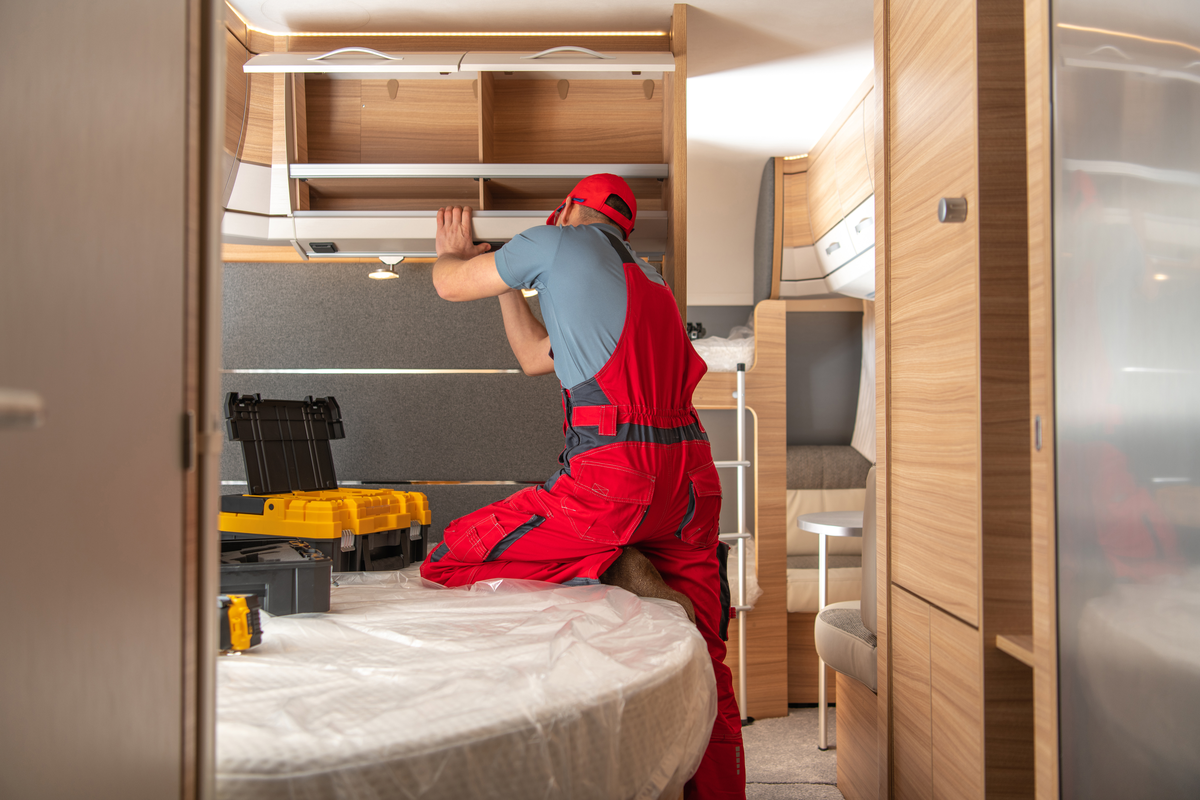 An RV technician repairing an overhead cabinet inside a travel trailer