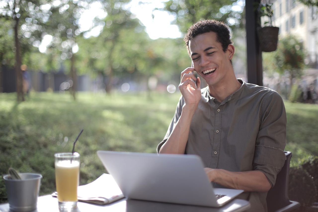 A smiling customer speaking on his phone beside a laptop at an outdoor cafe
