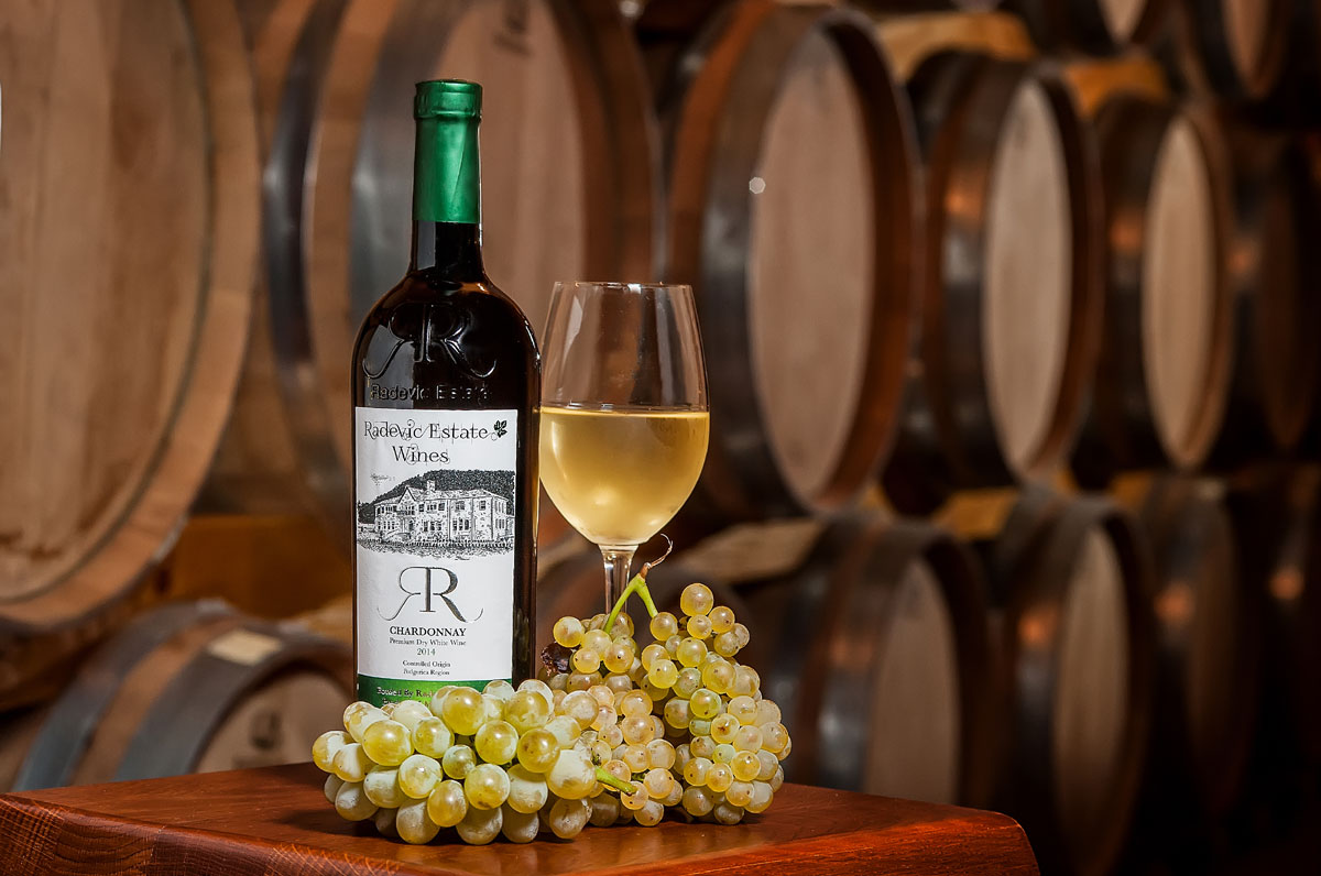 The flagship premium dry white wine from the family-operated Radević Estate winery in Rogami, Montenegro. Surrounded by their traditional barrels, the bottle is displayed alongside freshly picked Chardonnay grapes and a poured glass. The flagship premium dry white wine from the family-operated Radević Estate winery in Rogami, Montenegro. Surrounded by their traditional barrels, the bottle is displayed alongside freshly picked Chardonnay grapes and a poured glass.