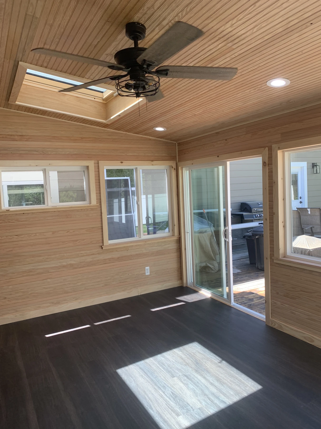 Wood-lined sunroom with a paneled ceiling and walls, windows, sliding glass doors and a dark floor.