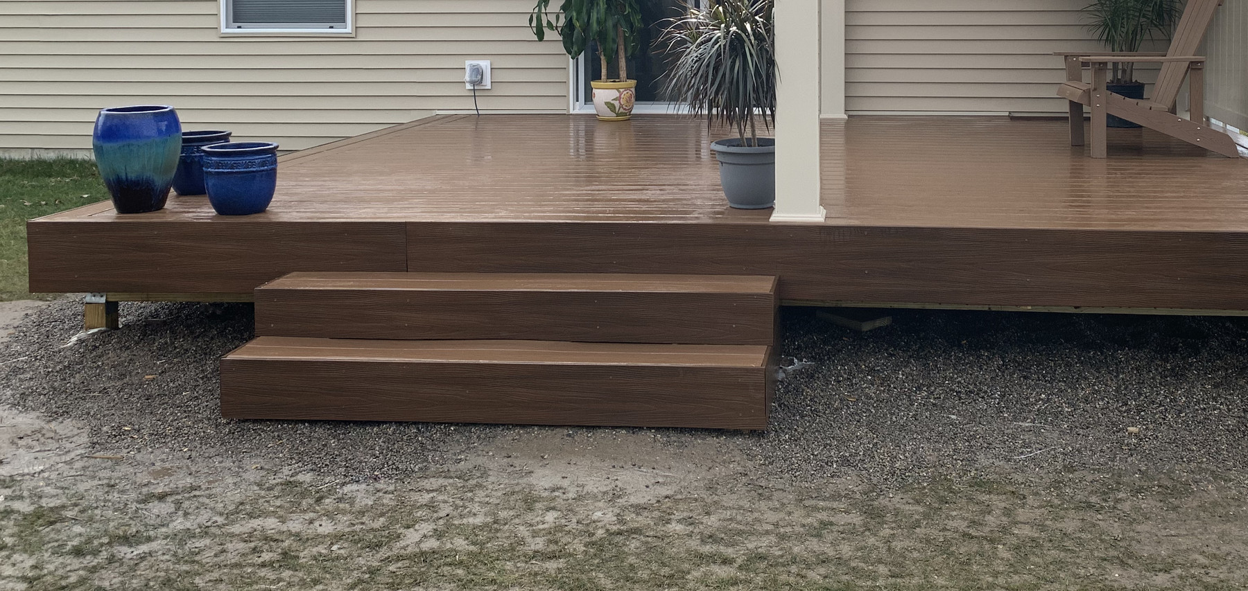 Front edge of a brown deck with broad steps, potted plants and the lower portions of white pergola posts.