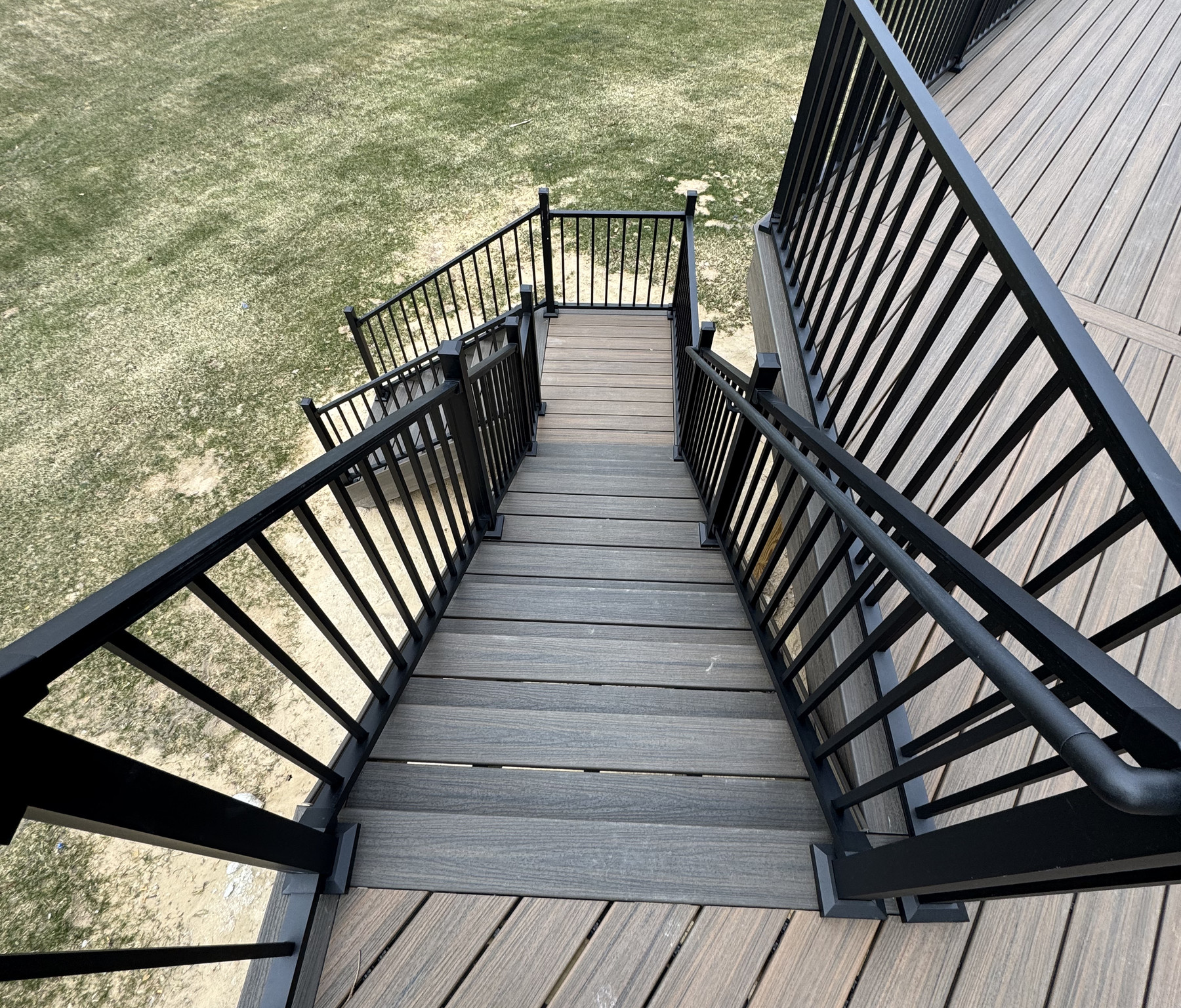View down gray deck steps toward a landing and the yard, with dark railings around the stairs and deck opening.