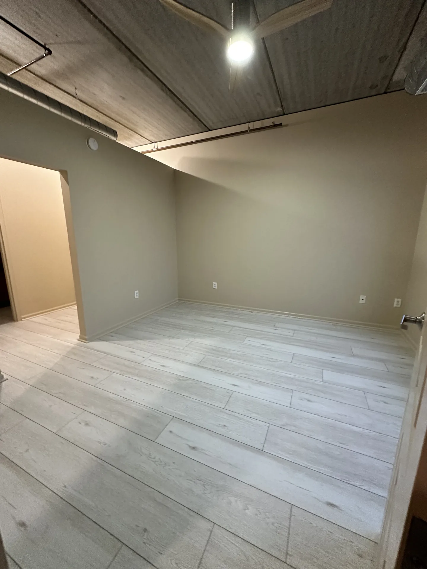 Interior room with pale gray floor planks, gray walls and an unfinished ceiling with exposed pipes.