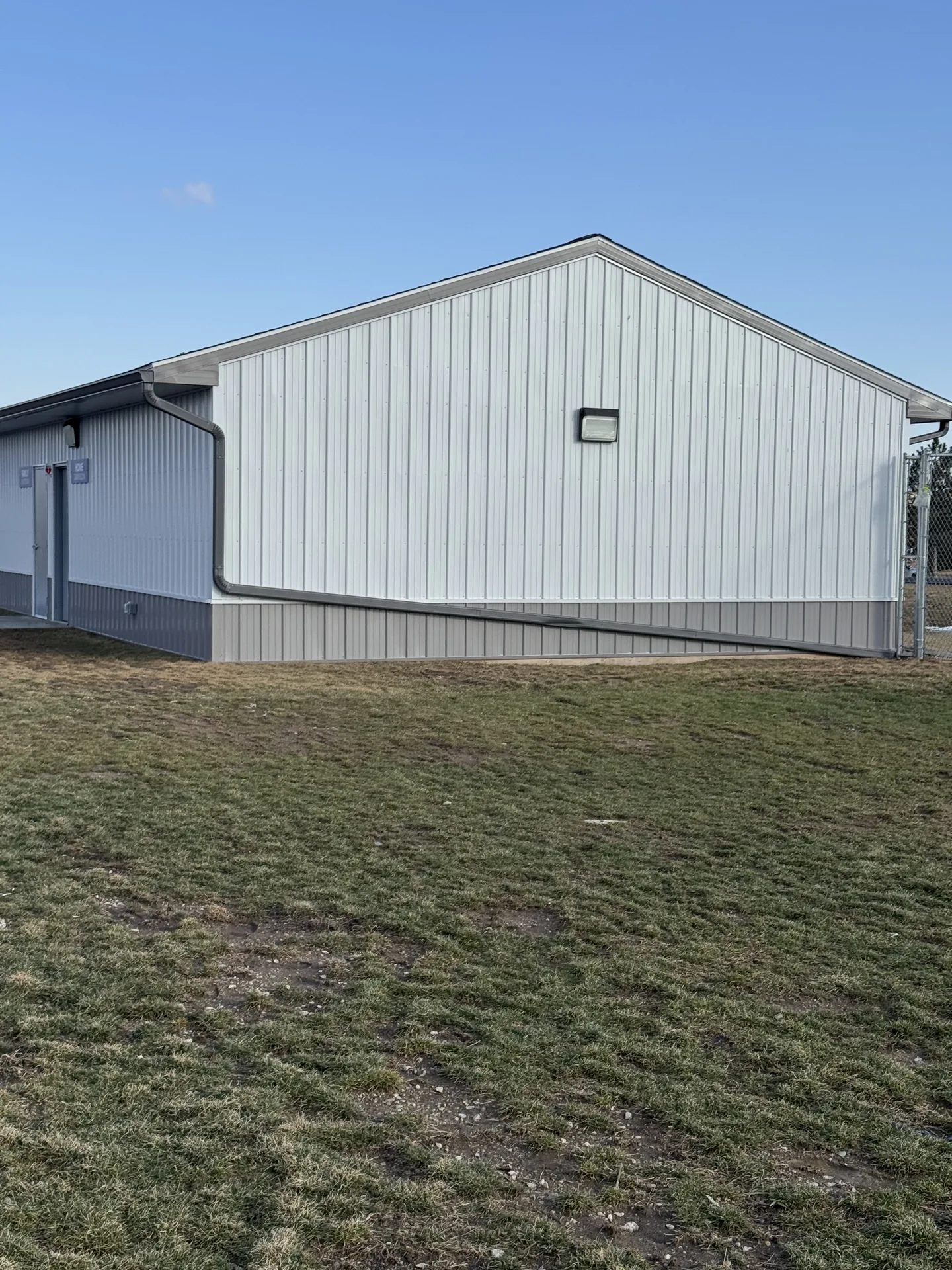 Light-gray barn exterior with a pitched roof, side entry doors and grass in the foreground.
