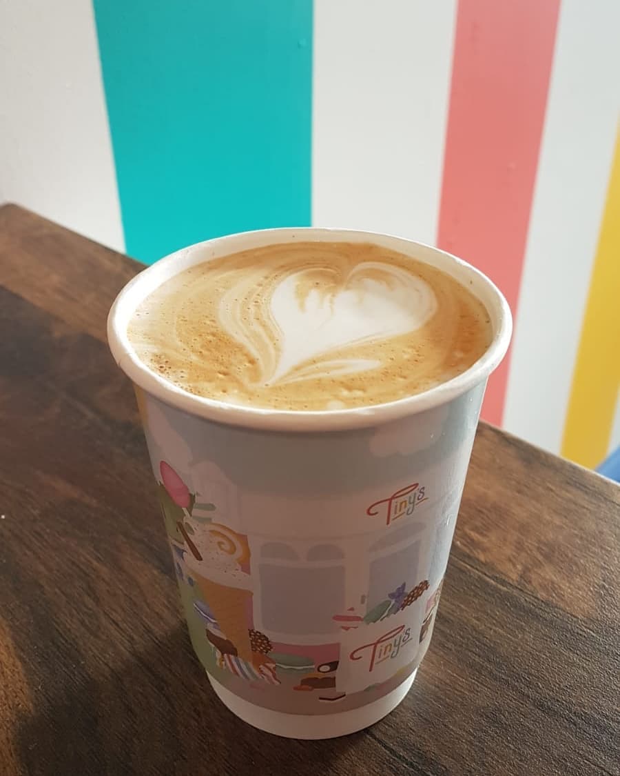 A latte in a takeaway cup against the colourful wall at Tiny's
