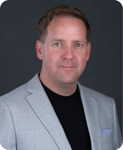 Professional headshot of Jay Lawrence, President & Co-Owner, wearing a light gray blazer and black shirt against a dark gray background.