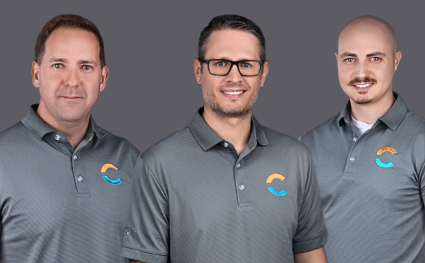 A group of four IT professionals wearing matching gray polo shirts with a company logo, posing for a team photo. They are standing against a neutral gray background, representing their IT support and managed services expertise.