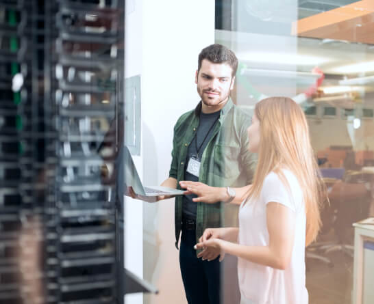 IT technician and a colleague discussing server management in a data center, with a laptop open for troubleshooting.