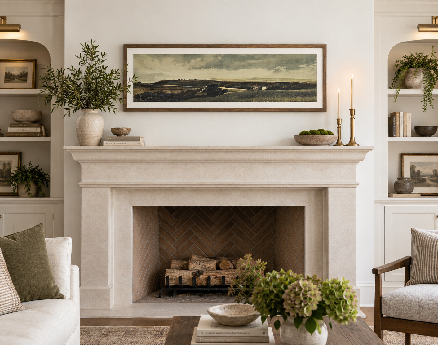 The same limestone fireplace with a long, narrow panoramic landscape in a natural wood frame above the mantel, stretching across roughly three-quarters of the shelf's width. A shallow bowl of moss and two brass candlesticks sit at one end.