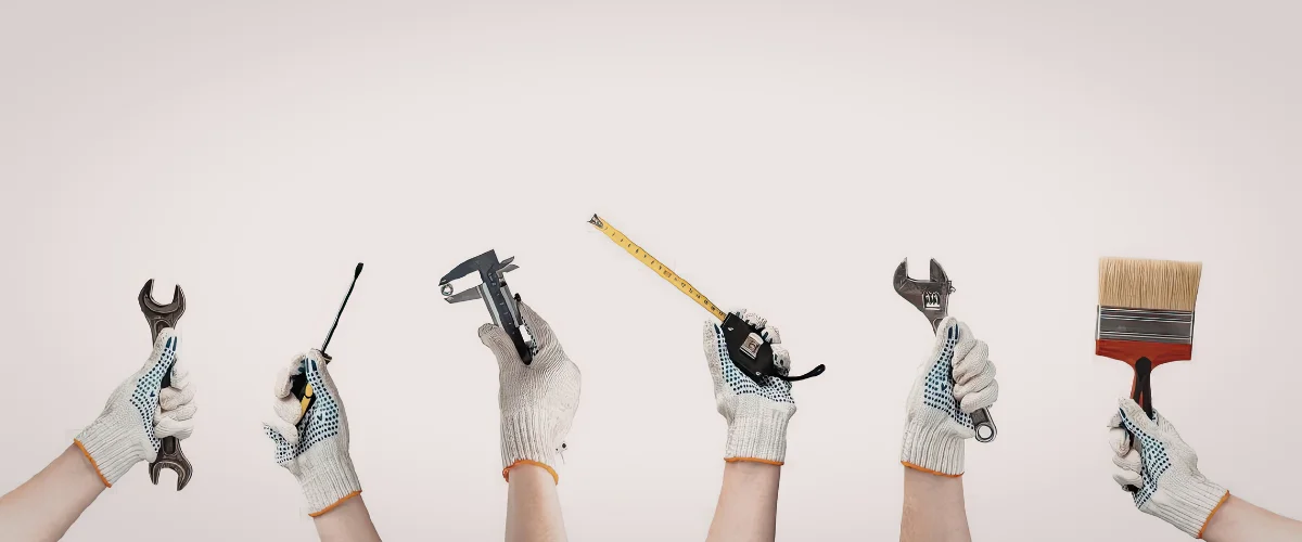 Six hands wearing white work gloves, each holding a different tool, against a light pink background.