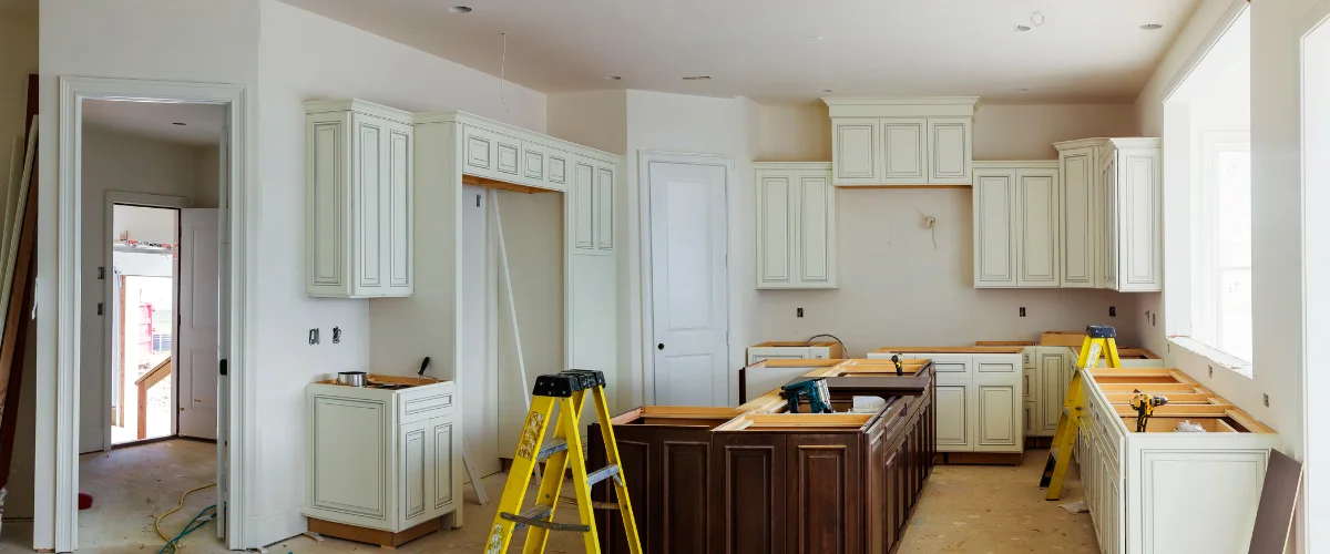 A new kitchen under construction with cream upper cabinets, brown base cabinets, and a yellow stepladder.