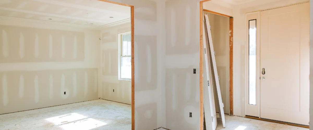 An interior view of a newly constructed house with unfinished drywall, exposed framing, and a partially installed front door.