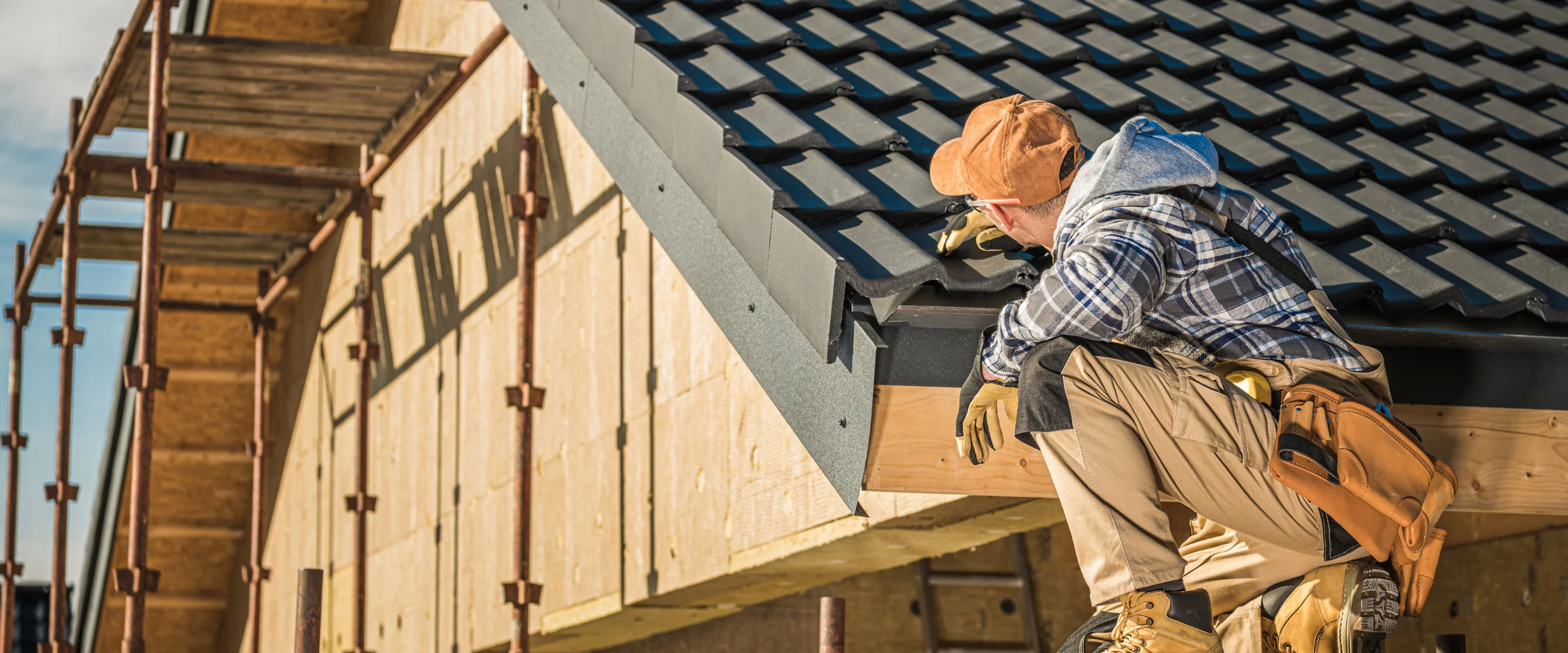 A construction worker in a plaid shirt and tan cap crouches on a roof installing dark gray tiles, with scaffolding visible left.