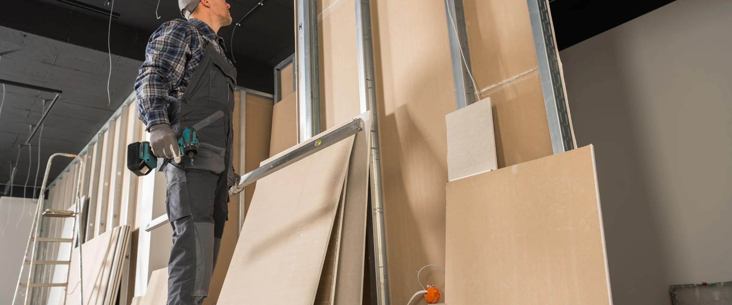 A construction worker in overalls holding a drill, looking up at an unfinished ceiling next to drywall panels and metal studs.
