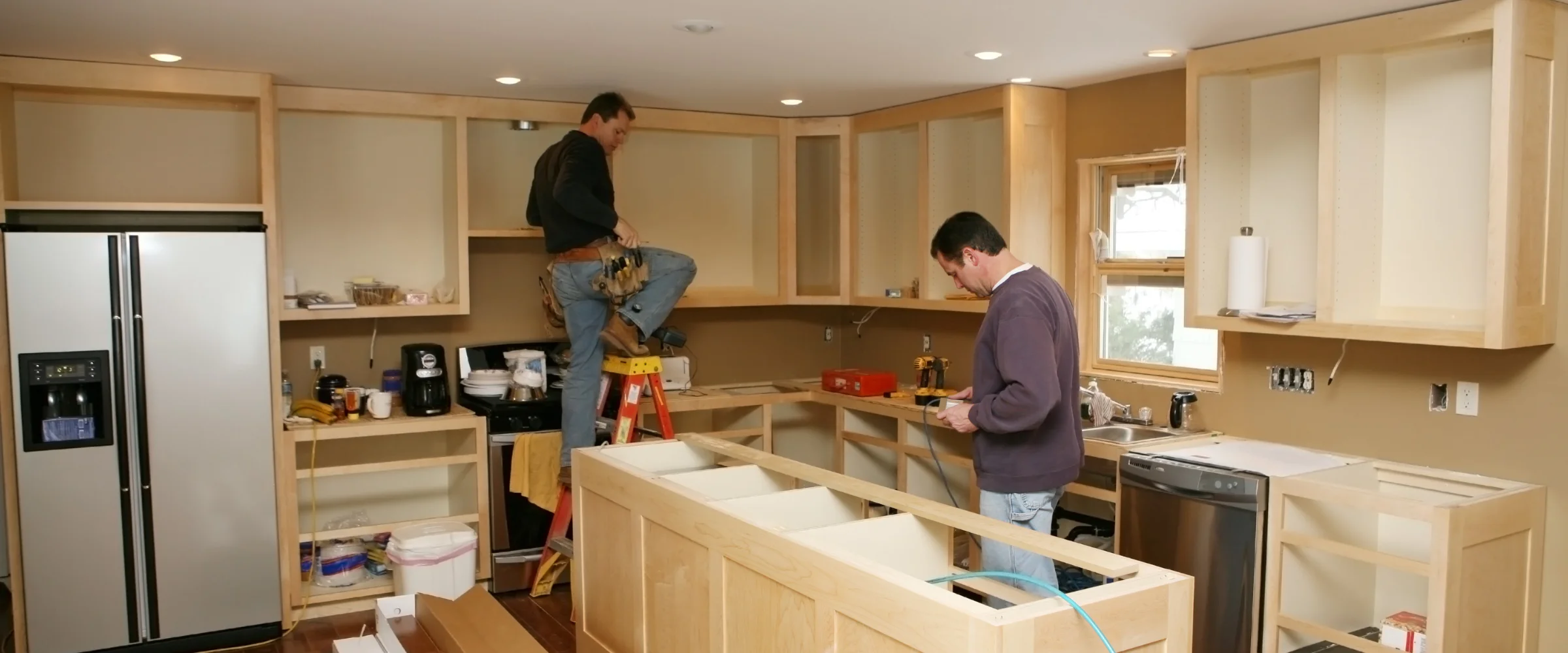 Two men installing light wood cabinets and island framing in a kitchen during renovation. Two men installing light wood cabinets and island framing in a kitchen during renovation.