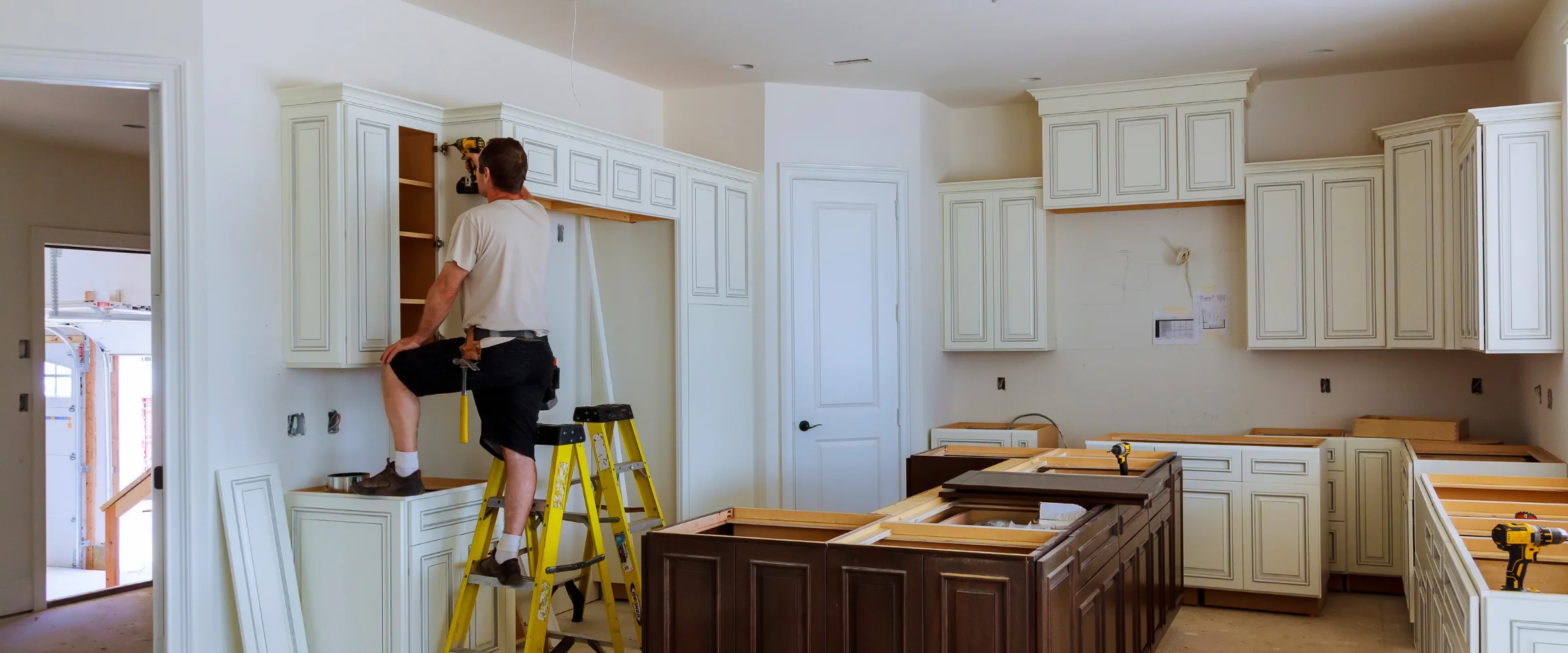 A man on a ladder installs white upper kitchen cabinets with a drill, while lower cabinets are set in an unfinished room. A man on a ladder installs white upper kitchen cabinets with a drill, while lower cabinets are set in an unfinished room.