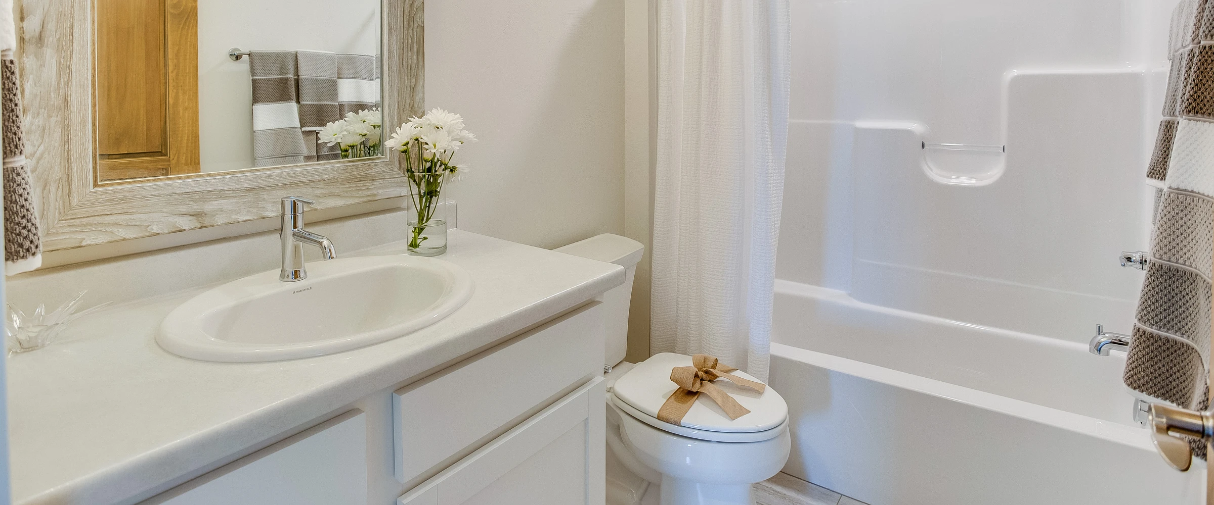 A bright, modern bathroom featuring a white vanity with sink, a framed mirror, a toilet with a burlap bow, and a white shower-tub combo.