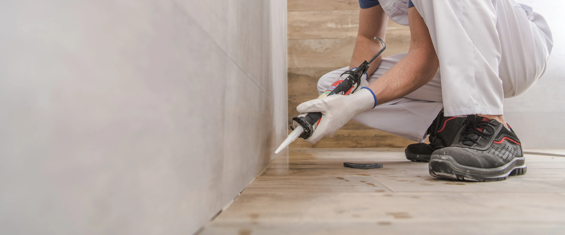 A gloved worker in white pants crouches, applying white sealant with a caulk gun along the joint between a light tile wall and a wood-look floor.