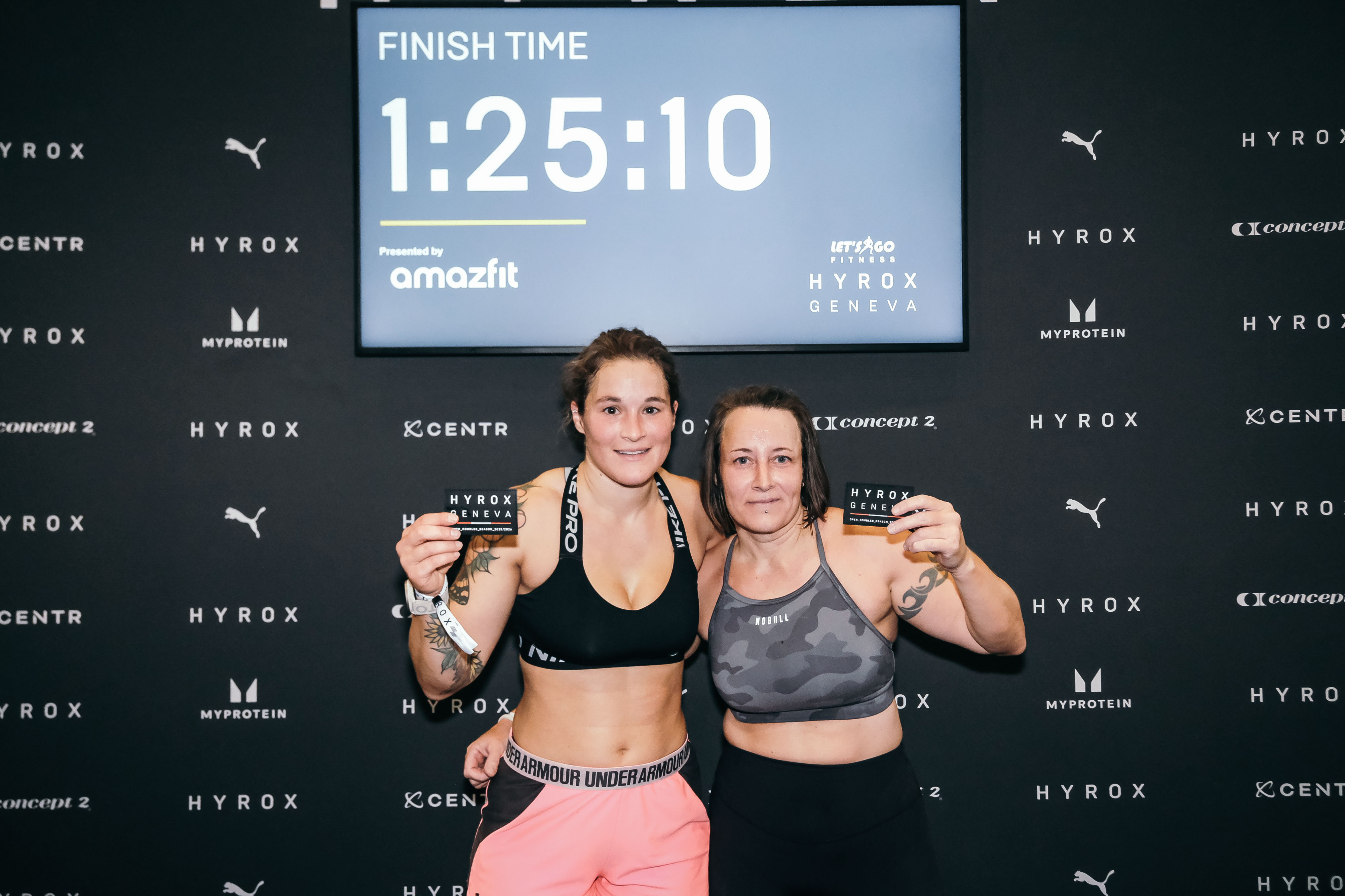 Deux participantes après leur Hyrox Genève, tenant leurs cartes de finisher devant l'écran affichant leur temps