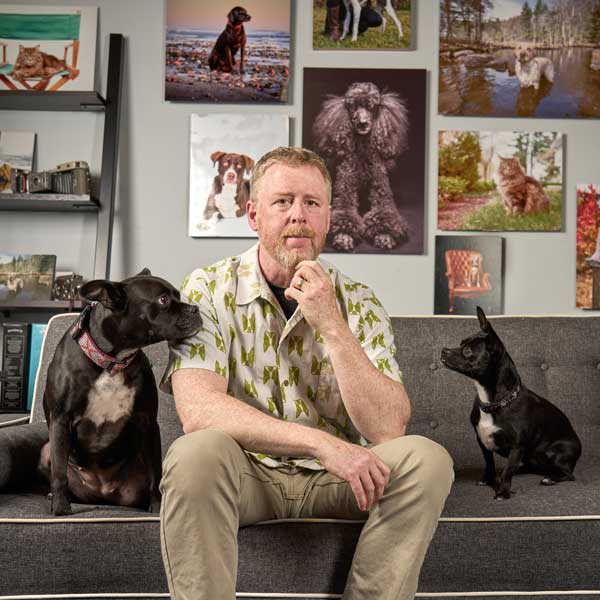 Fred Levy, sitting on a couch in his studio with photos behind him and his two dogs on either side.