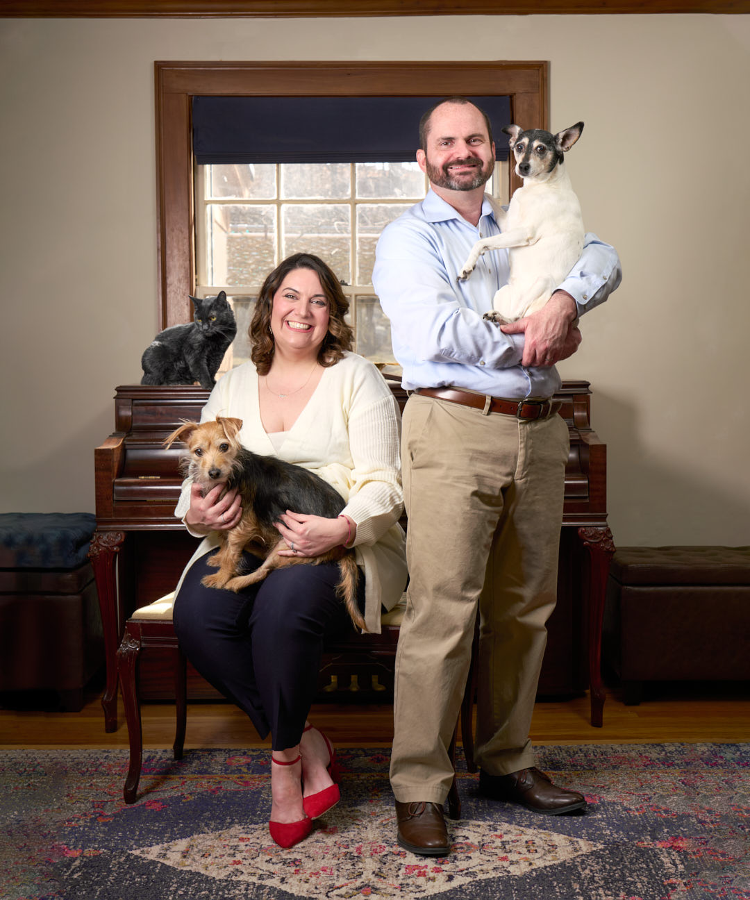 Couple in front of a piano, each holding a dog with a cat sitting on the piano