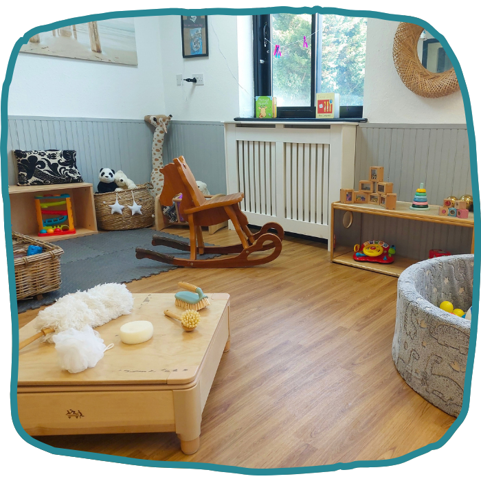 A bright playroom with a wooden rocking chair, low tables and shelves of toys, stuffed animals, a rug, and a window.