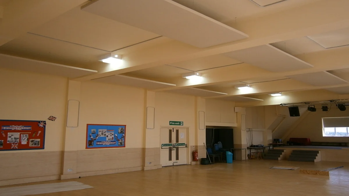 Rows of white suspended acoustic panels above a wooden-floored hall with a small stage.
